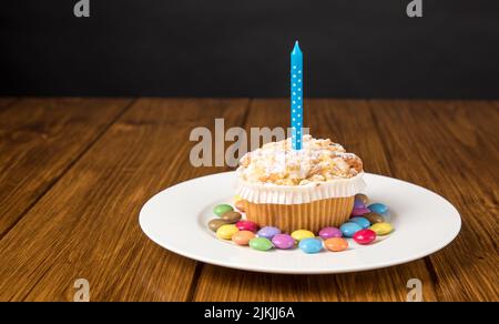 Ein Teller mit Muffin mit brennender Kerze und bunten Süßigkeiten auf einem Holztisch in der Nähe der grauen Wand Stockfoto