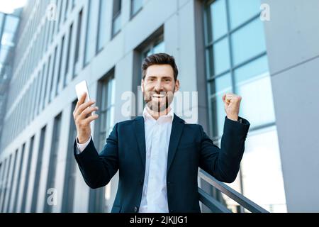 Froher kaukasischer Tausendjähriger Mann mit Bart im Anzug mit Smartphone freut sich über den Sieg und hebt die Hände hoch Stockfoto