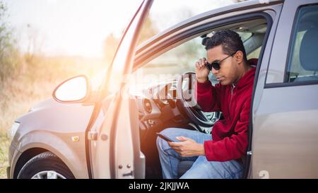 Hübscher junger Hispanic, der sein Auto benutzt und sein Smartphone anschaut Stockfoto