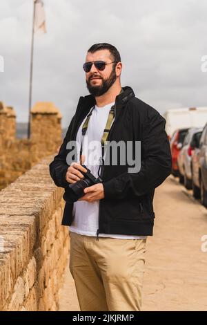 Eine vertikale Aufnahme eines jungen bärtigen Touristen-Mannes in Sonnenbrille mit einer Kamera auf einer Brücke Stockfoto