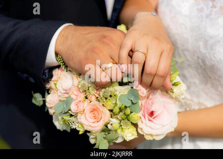 Eine Nahaufnahme der Hände der Braut und des Bräutigams, die Hochzeitsbänder auf einem Blumenstrauß tragen Stockfoto