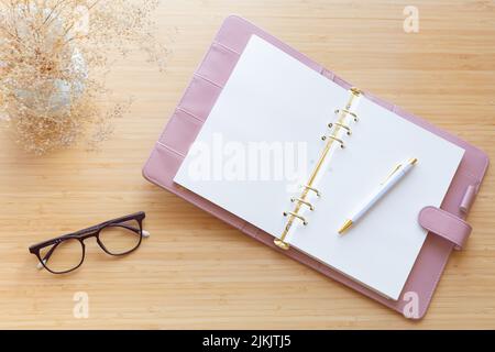 Flach liegend, Draufsicht auf einen pastellrosa Planer, getrockneter Blumenstrauß, Gläser und Papeterie auf einer Holzoberfläche. Schicker Arbeitsplatz, Bloggen. Stockfoto