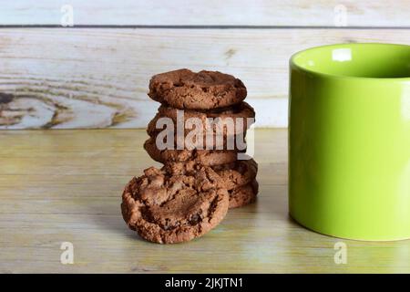 Eine grüne Tasse Tee neben gestapelten Schokoladenkeksen auf einem Holztisch-Hintergrund Stockfoto