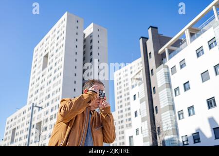 Ein älterer Mann fotografiert mit einer alten Filmkamera in der Stadt mit Gebäuden im Hintergrund. Stockfoto