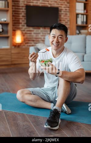 Gesundes Lifestyle-Konzept. Ein glücklicher Mann mittleren Alters, der Gemüsesalat isst, auf einer Yogamatte sitzt und vor der Kamera lächelt Stockfoto
