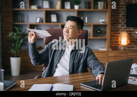 Fröhlicher, gutaussehender asiatischer Geschäftsmann mittleren Alters spielt mit einem Papierflugzeug am Arbeitsplatz am Laptop zu Hause Stockfoto