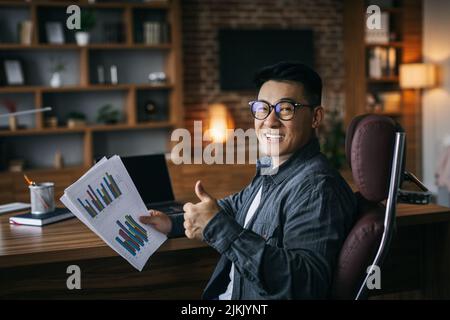 Glücklicher Erwachsener asiatischer Geschäftsmann in einer Brille, der mit Diagrammen arbeitet und Daumen zeigt und an ein Online-Projekt denkt Stockfoto
