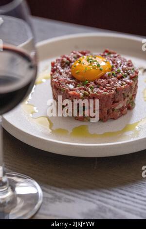 Ein vertikaler selektiver Fokus aus einer Steak-Tartare mit Eigelb auf einem weißen Teller und einem Glas Rotwein Stockfoto