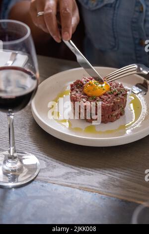 Ein vertikaler selektiver Fokus aus einer Steak-Tartare mit Eigelb auf einem weißen Teller und einem Glas Rotwein Stockfoto