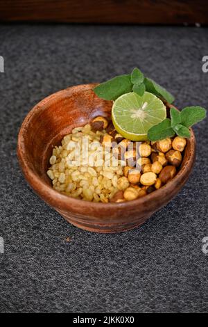 Nahaufnahme der gebratenen gelben Gramm Split Linsen mit schwarzem Gramm und in Scheiben geschnittene Zitrone, Minze in der roten Holzschale über unscharfem grauen Hintergrund. Stockfoto