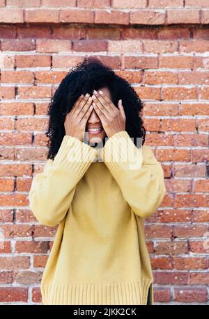 Eine Afrikanerin aus Spanien in gelbem Pullover, der das Gesicht mit Händen mit Backsteinmauer auf dem Hintergrund bedeckt Stockfoto