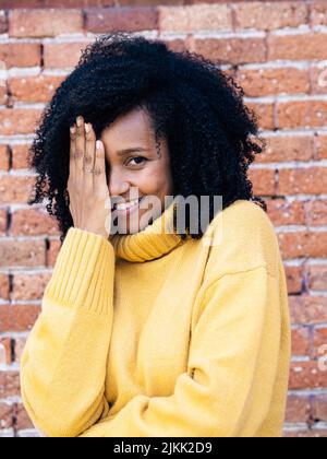 Eine Afrikanerin aus Spanien in gelbem Pullover, der das Gesicht mit der Hand mit einer Backsteinwand auf dem Hintergrund bedeckt Stockfoto