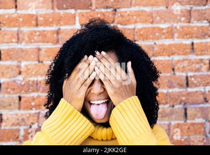 Eine Afrikanerin aus Spanien in gelbem Pullover, der das Gesicht mit Händen mit Backsteinmauer auf dem Hintergrund bedeckt Stockfoto