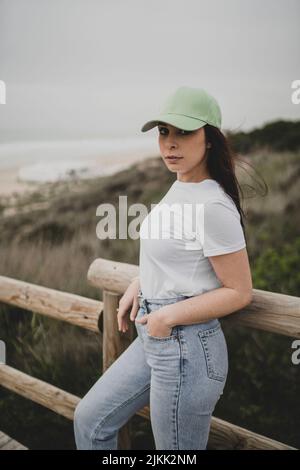 Eine niedliche junge kaukasische Dame, die eine Mütze, ein T-Shirt und eine Jeans trägt und in einem ländlichen Gebiet posiert Stockfoto