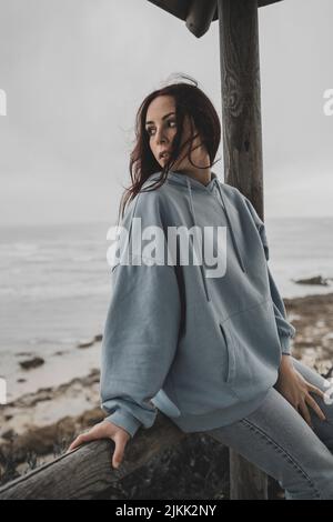 Eine niedliche junge kaukasische Dame mit Kapuzenpullover und Jeans sitzt in einem Pavillon am Meer Stockfoto