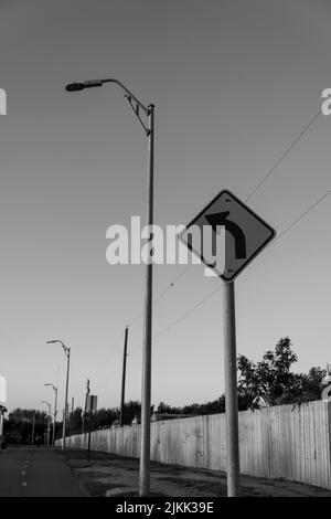 Eine Graustufenaufnahme eines Verkehrszeichens mit einer Linkskurve vor einem Straßenpfosten in der Straße neben einer Straßenlaterne Stockfoto
