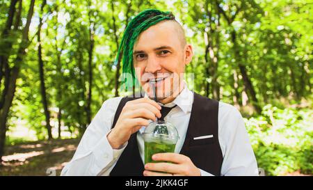 Junger Mann mit Dreadlocks trinkt erfrischende Limonade im Waldpark. Stilvoller Hipster im Business-Anzug mit grüner Frisur und alkoholfreien Getränken Stockfoto