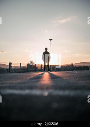 Eine vertikale Aufnahme aus dem niedrigen Winkel einer Rückansicht eines Mannes, der bei Sonnenuntergang auf der Straße steht Stockfoto