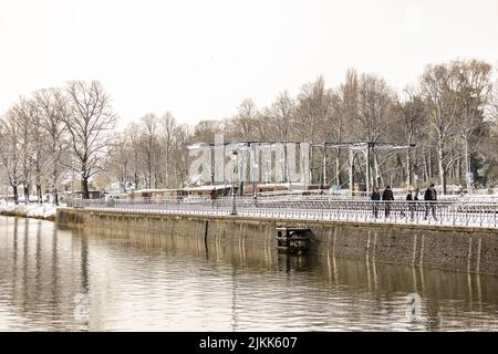 Menschen, die in der Wintersaison im Schnee den Leidsche Rijn Kanal entlang schlürfen, mit Stahlbrücke im Hintergrund. Stockfoto