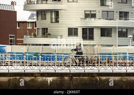 Frau, die auf einem verschneiten Kai mit einem Fahrrad unterwegs ist und im Hintergrund einen modernen, modernen Wohnturm hat. Niederländische Wetterbedingungen und Transport Stockfoto