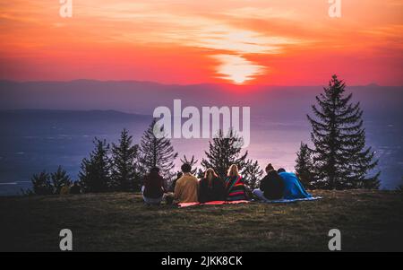 Eine Rückansicht einer Gruppe von Menschen, die am Seeufer auf einem faszinierenden Sonnenuntergang am Himmel sitzen Stockfoto