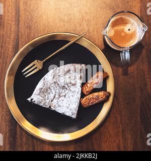 Eine Draufsicht auf ein Stück Kuchen und zwei Kekse auf dem schwarz-goldenen Teller mit einer Tasse Kaffee Stockfoto
