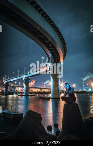Ein Mensch mit beleuchteter Maske sitzt auf einem Felsen im Hintergrund der Yokkaichi-Brücke in der Nacht Stockfoto