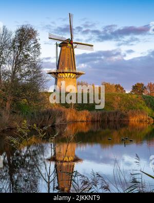 Eine vertikale Aufnahme einer alten Windmühle, die an einem bewölkten Tag in einem Flusswasser reflektiert wird Stockfoto