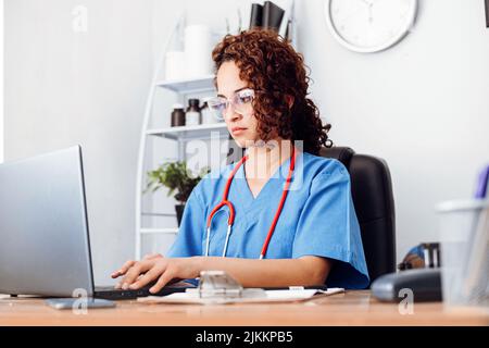 Gesundheitspersonal, das an einem Computer arbeitet, latina Ärztin mit Brille, die in ihrem Büro einen Laptop benutzt. Stockfoto