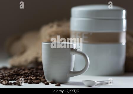 Ein flacher Fokus einer Tasse heißen Kaffees mit Kaffeebohnen und weißem Behälter im Hintergrund Stockfoto