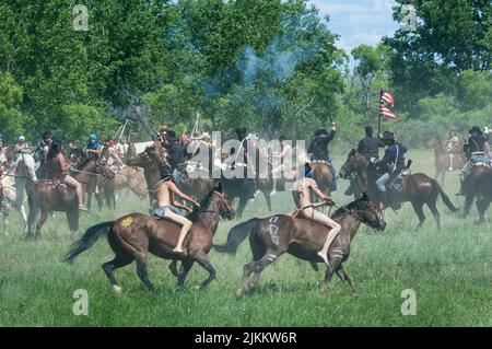 US-Soldaten in Kavallerieuniformen reiten zu Pferd und schießen Pistolen, während sie während der Little Bighorn Nachstellung A gegen indische Sioux-Braves kämpfen Stockfoto