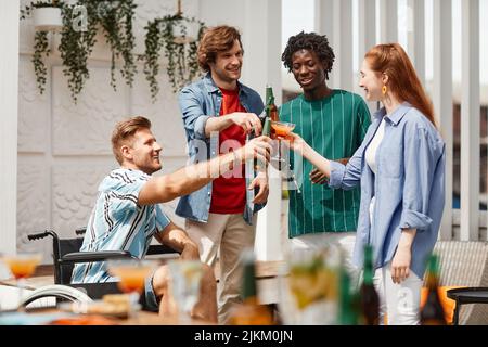 Diverse Freunde, darunter Rollstuhlfahrer, die mit Getränken toasten, während sie die Party auf der von Sonnenlicht beleuchteten Außenterrasse genießen Stockfoto
