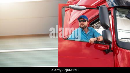 Kaukasischer Semi-Truck-Fahrer Trägt Eine Sonnenbrille In Der Fahrzeugkabine. Vorbereitung auf einen langen Weg. Thema Schwertransportindustrie. Stockfoto