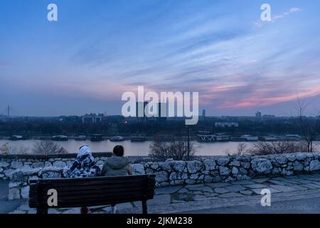 Eine Rückansicht eines Paares auf der Bank in der Nähe des Seeufers auf einem Sonnenuntergang Himmel Hintergrund sitzen Stockfoto