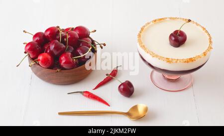 Dunkle und weiße Schokoladenmousse oder Panna Cotta mit Chilikirschgelee in einem Glas mit Holzschüssel mit reifen Kirschen auf weißem Holztisch. Hoher Winkel Stockfoto