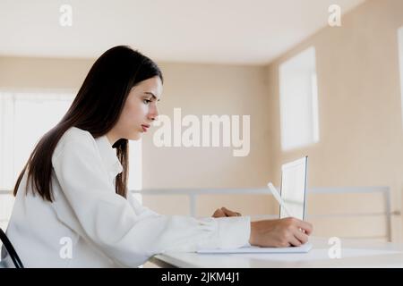 Fokussierte kaukasische junge Geschäftsfrau, die am Schreibtisch sitzt, am Computer liest und sich an ihrem weißen Notizbuch Notizen macht. Studentin mit pc, die wichtige Informationen mit Bleistift in Notizblock notiert Stockfoto