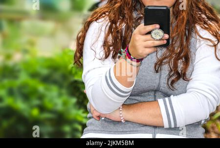 Nahaufnahme einer Frau, die ein Handy hält und liest, in einem schwarzen Etui mit Griff auf dem Rücken - langes Haar mit niedlichen Armbändern und Kapuzenoberteil - Casua Stockfoto