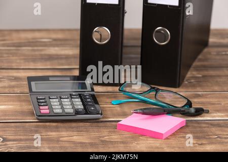 Eine Nahaufnahme von Stift auf pinkfarbenen Haftnotizpapieren, Taschenrechner, blauen Gläsern und schwarzen Papiermappen auf einem Holzhintergrund Stockfoto