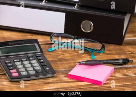 Eine Nahaufnahme von Stift auf pinkfarbenen Haftnotizpapieren, Taschenrechner, blauen Gläsern und schwarzem Papierordner auf einem Holzhintergrund Stockfoto