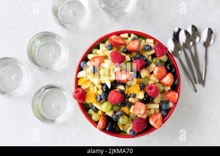 Eine Draufsicht auf bunten Obstsalat in einer Schüssel mit Glascops und Löffeln auf weißem Hintergrund Stockfoto