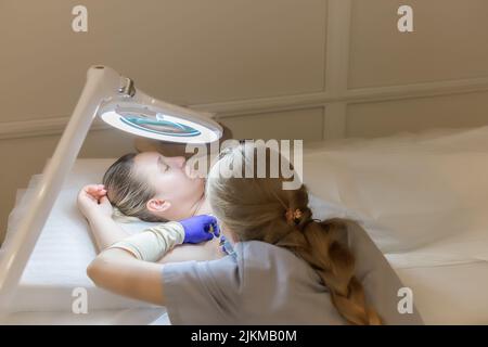 Der Meister der Elektrolyse in blauen sterilen Handschuhen führt das Verfahren zur Entfernung unerwünschter Haare mittels Elektrolyse durch Stockfoto