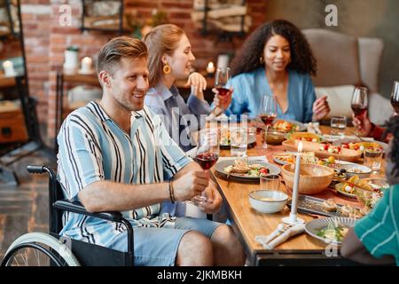 Porträt eines lächelnden jungen Mannes im Rollstuhl, der mit Freunden eine Dinner-Party genießt und Weinglas und einen Kopierraum hält Stockfoto