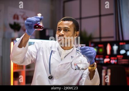 Yong afrikanischer Wissenschaftler, der mit Reagenzgläsern mit Labormantel arbeitet, arbeitet im Labor, während er die biochemische Probe in Reagenzgläsern und wissenschaftlichen Instrumenten untersucht. Stockfoto