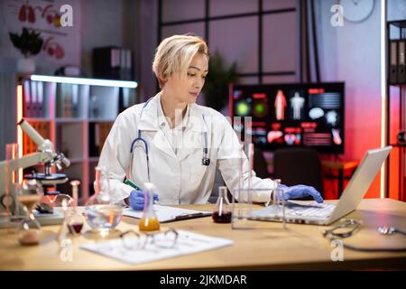 Wissenschaftlerin, die im Labor an einem Laptop arbeitet. Die junge kaukasische Forscherleiterin führt Untersuchungen mit Reagenzgläsern durch, während sie Forschungsergebnisse schreibt. Stockfoto