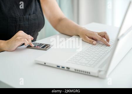 Frau sitzt am Laptop und macht Berechnungen auf dem Rechner. Nahaufnahme von Händen, Taschenrechner und Laptop. Konzept der Finanz- und Projektberechnungen und Rechnungslegung. Stockfoto