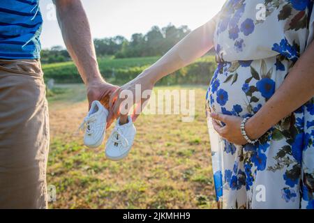 Ein Paar werdende Eltern hält Babyschuhe, die Mutter berührt ihren schwanger werdenden Bauch, das wartende Paar Stockfoto