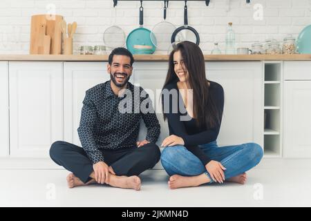 Das Paar sitzt barfuß auf dem Boden an der Küchentheke, entspannt lächelnd und lachend und blickt auf die Kamera. Innenaufnahme. Hochwertige Fotos Stockfoto