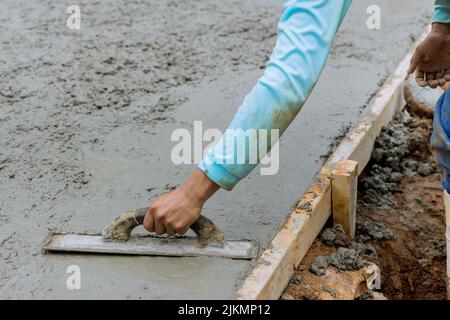 Der Mason-Arbeiter hält eine Stahlkelle und glättet neue Gehwege auf nassem, frisch gegossenen Beton Stockfoto