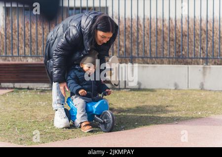 Die junge, schöne Mutter kümmert sich um sein kleines, niedliches afrikanisches Kind und lehrt es, in einem Hintergrund in der Nähe des Zauns ein Baby-Fahrrad zu fahren. Hochwertige Fotos Stockfoto