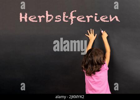 Mädchen schreibt auf einer Tafel den deutschen Text Herbstferien, in englischer Sprache Herbstferien Stockfoto